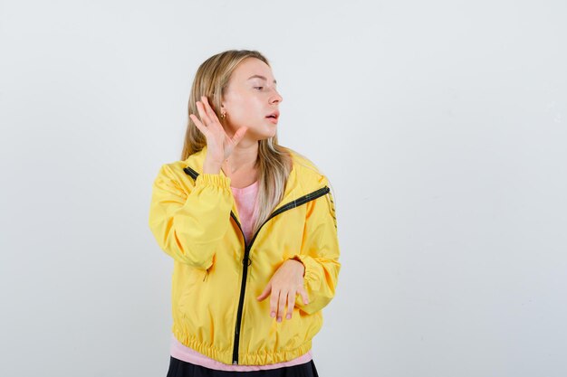 Blondes Mädchen, das Händchen in der Nähe des Ohrs hält, um etwas in rosa T-Shirt und gelber Jacke zu hören und fokussiert zu schauen