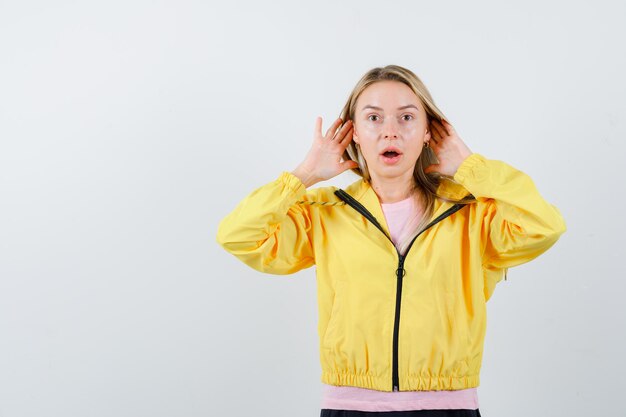 Blondes Mädchen, das Händchen in der Nähe des Ohrs hält, um etwas in rosa T-Shirt und gelber Jacke zu hören und überrascht zu schauen.