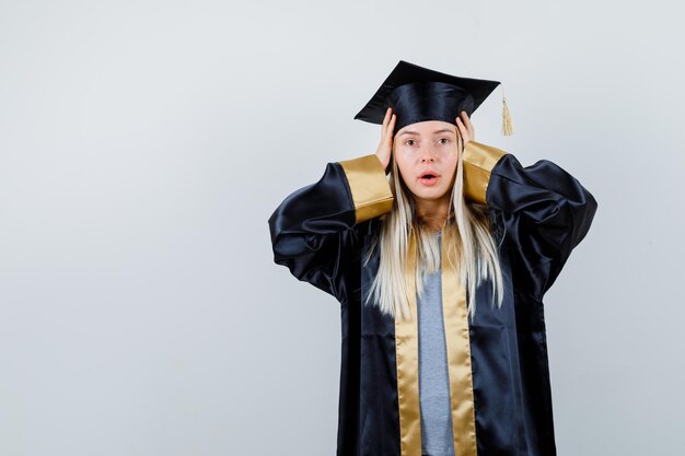 Blondes Mädchen, das Hände in Abschlusskleid und Mütze aufsetzt und überrascht aussieht