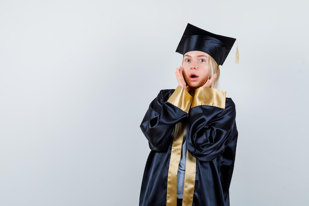 Blondes Mädchen, das Hände in der Nähe des Gesichts hält, den Mund in Abschlusskleid und Mütze öffnet und überrascht aussieht