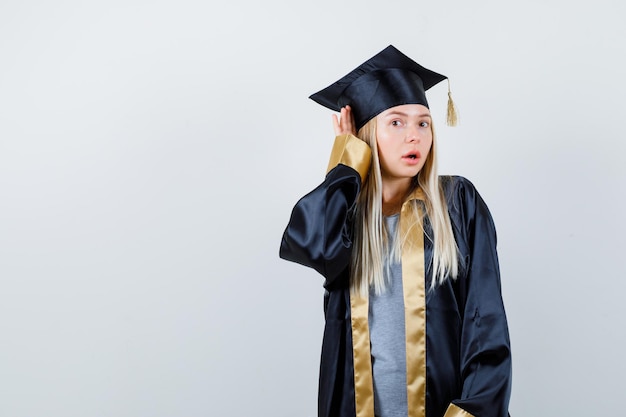 Kostenloses Foto blondes mädchen, das hände in der nähe des ohres hält, um in abschlusskleid und mütze zu hören und überrascht zu schauen.