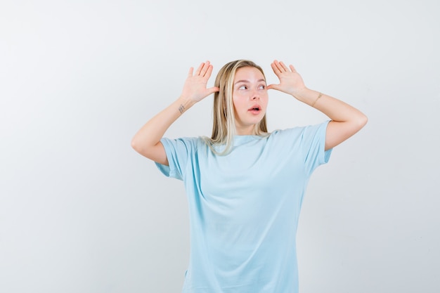 Blondes Mädchen, das Hände nahe Kopf hält, weg im blauen T-Shirt schaut und niedlich schaut. Vorderansicht.