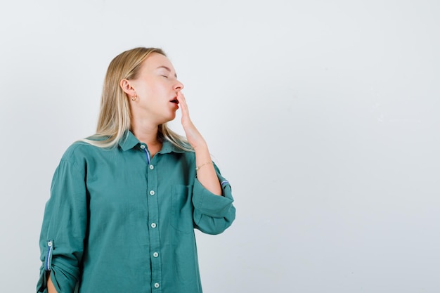 Blondes Mädchen, das Hand auf den Mund legt, in grüner Bluse gähnt und schläfrig aussieht looking