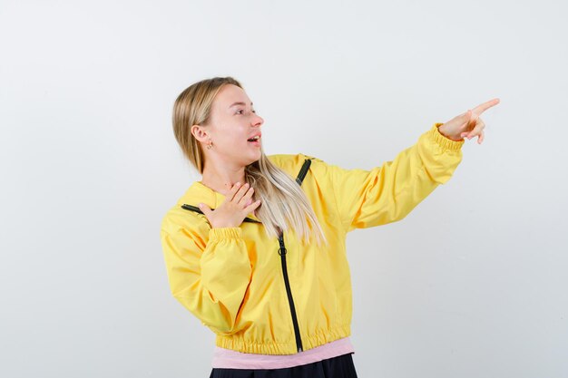 Blondes Mädchen, das Hand auf die Brust legt, in rosa T-Shirt und gelber Jacke nach rechts zeigt und überrascht aussieht.