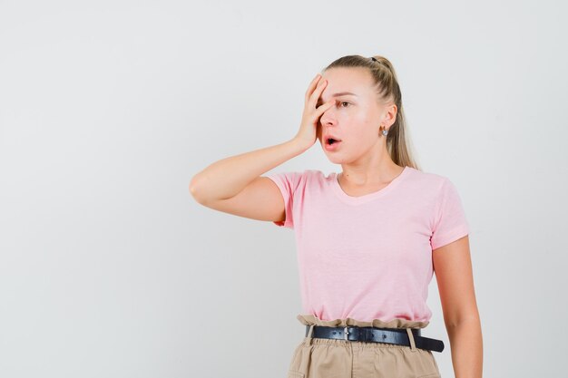 Blondes Mädchen, das Hand auf Gesicht in T-Shirt, Hose hält und deprimiert aussieht. Vorderansicht.