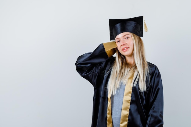 Blondes Mädchen, das Hand hinter dem Kopf in Abschlusskleid und Mütze streckt und süß aussieht