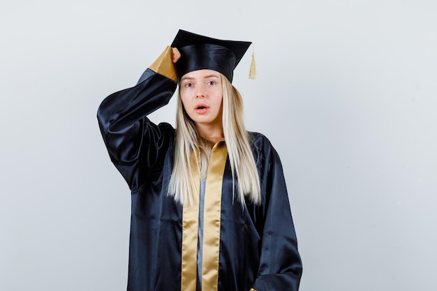 Blondes Mädchen, das Hand in Abschlusskleid und Mütze auf den Kopf legt und süß aussieht.