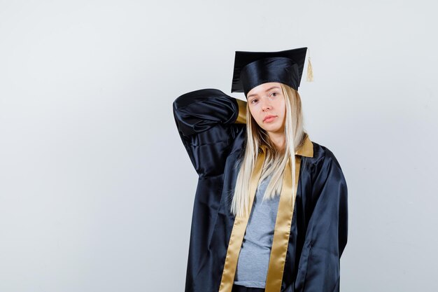 Blondes Mädchen, das Hand in Absolventenuniform am Hals hält und süß aussieht