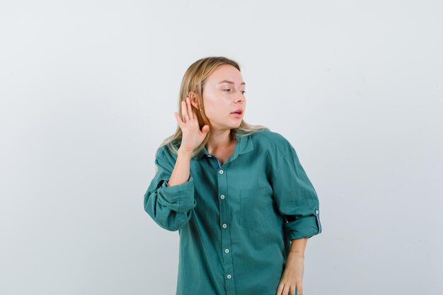 Blondes Mädchen, das Hand in der Nähe des Ohrs hält, um etwas in grüner Bluse zu hören und konzentriert aussieht