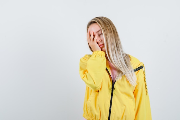 Blondes Mädchen, das Hand in gelber Jacke im Gesicht hält und erschöpft aussieht