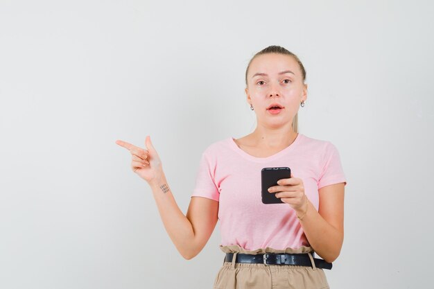 Blondes Mädchen, das Handy hält und in T-Shirt, Hosenvorderansicht beiseite zeigt.