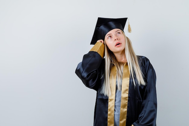 Blondes Mädchen, das in denkender Pose in Abschlusskleid und Mütze steht und nachdenklich aussieht