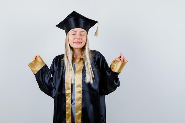 Blondes Mädchen, das in meditierender Pose in Abschlusskleid und Mütze steht und ruhig aussieht.