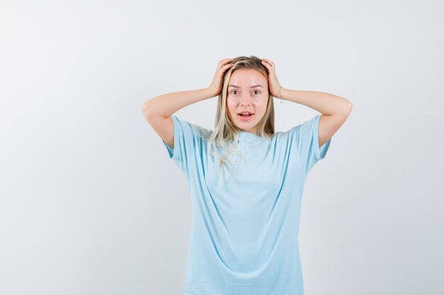 Blondes Mädchen, das Kopf mit Händen im blauen T-Shirt umklammert und hübsch aussieht. Vorderansicht.