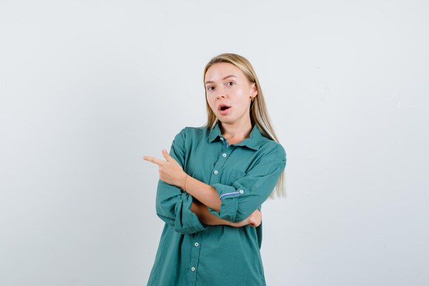 Blondes Mädchen, das mit dem Zeigefinger in der grünen Bluse nach links zeigt und überrascht schaut.