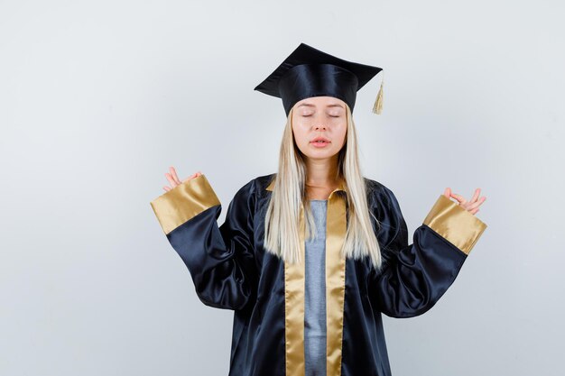 Blondes Mädchen, das mit geschlossenen Augen in Absolventenuniform meditiert und hoffnungsvoll aussieht