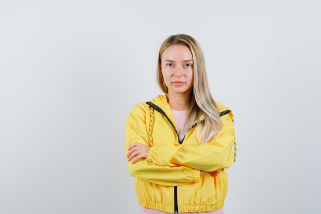 Blondes Mädchen, das mit verschränkten Armen in gelber Jacke steht und selbstbewusst aussieht.