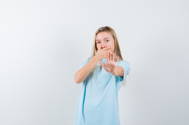 Blondes Mädchen, das Mund mit Hand bedeckt, Stoppschild im blauen T-Shirt zeigt und überrascht, Vorderansicht schaut.