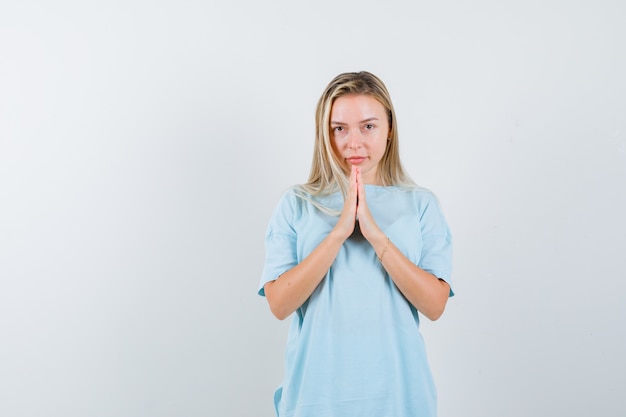 Blondes Mädchen, das namaste Geste im blauen T-Shirt zeigt und hübsch aussieht. Vorderansicht.