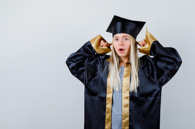 Blondes Mädchen, das Ohren mit Zeigefingern in Abschlusskleid und Mütze steckt und überrascht aussieht