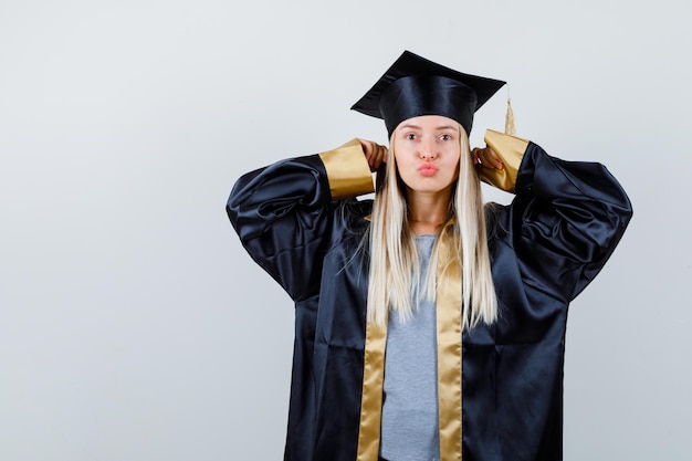 Blondes Mädchen, das Ohren mit Zeigefingern verstopft, Küsse in Abschlusskleid und Mütze sendet und glücklich aussieht