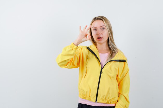 Blondes Mädchen, das okayzeichen in rosa T-Shirt und gelber Jacke zeigt und glücklich aussieht.