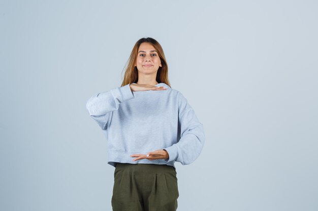 Blondes Mädchen, das sich die Hände ausdehnt, als etwas in olivgrünem blauem Sweatshirt und Hosen hält und fröhlich aussieht, Vorderansicht.