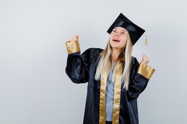 Blondes Mädchen, das Siegergeste im Abschlusskleid und in der Kappe zeigt und glücklich aussieht.