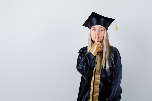 Blondes Mädchen, das Stillegeste in Abschlusskleid und Mütze zeigt und süß aussieht.