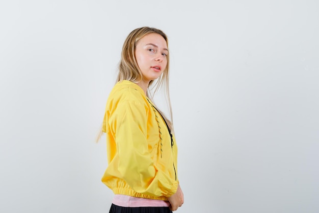 Blondes Mädchen, das über die Schulter schaut, während es in rosa T-Shirt und gelber Jacke in die Kamera posiert und verführerisch aussieht.