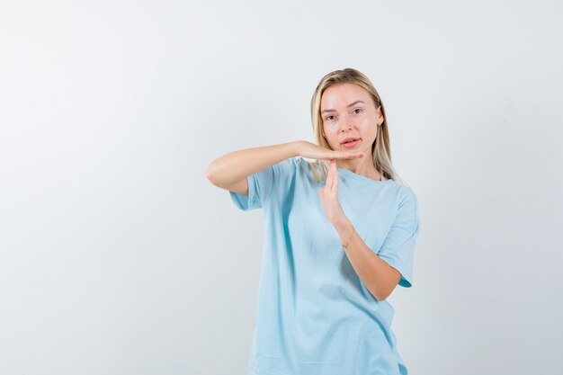 Blondes Mädchen, das Zeitpausengeste im blauen T-Shirt zeigt und hübsch aussieht. Vorderansicht.