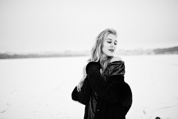 Blondes Mädchen der Eleganz im Pelzmantel und im roten Abendkleid stellte am verschneiten Wintertag auf