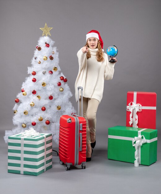 Blondes Mädchen der Vorderansicht mit Weihnachtsmütze, die Globus nahe rotem Koffer hält