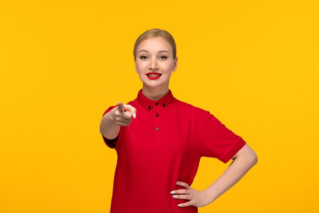 Blondes Mädchen des roten Hemdtages, das in einem roten Hemd auf einem gelben Hintergrund lächelt und nach vorne zeigt