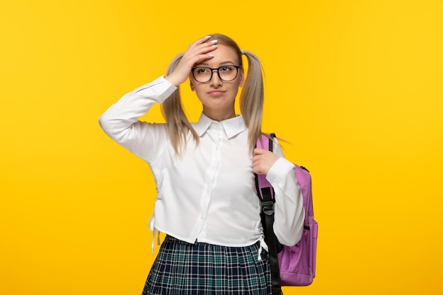 Blondes Mädchen des Weltbuchtages, das die Stirn mit Rucksack auf gelbem Hintergrund berührt