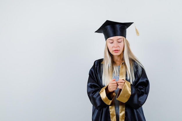 Blondes Mädchen gestikuliert wie das Spielen mit dem Handy in Abschlusskleid und Mütze und schaut fokussiert