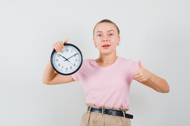 Blondes Mädchen hält Wanduhr, zeigt Daumen oben in T-Shirt, Hose und schaut lustig, Vorderansicht.