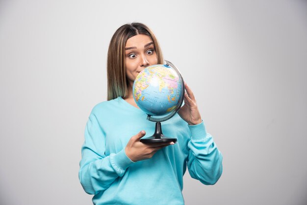 Blondes Mädchen im blauen Sweatshirt, das einen Globus hält, Standort errät und Spaß hat.