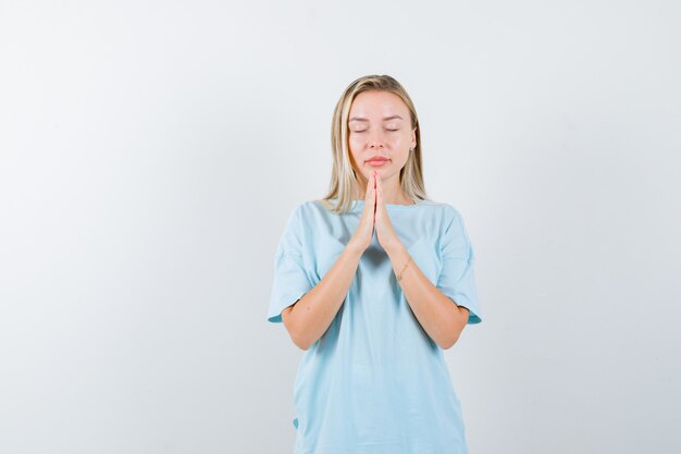 Blondes Mädchen im blauen T-Shirt, das Hände in betender Position fasst und ruhige Vorderansicht schaut.