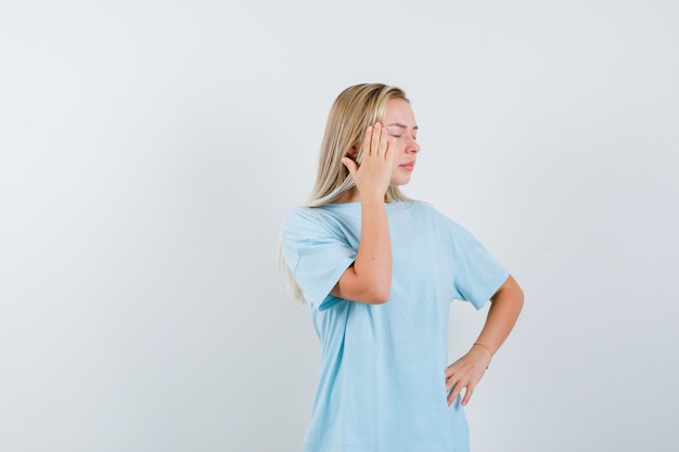 Blondes Mädchen im blauen T-Shirt, das Hand auf Schläfe setzt, Hand auf Taille hält und müde, Vorderansicht schaut.