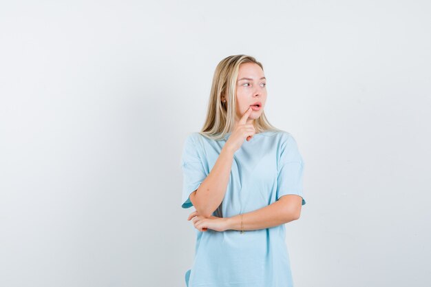 Blondes Mädchen im blauen T-Shirt, das in der denkenden Haltung steht, Zeigefinger nahe Mund setzt und nachdenklich schaut, Vorderansicht.