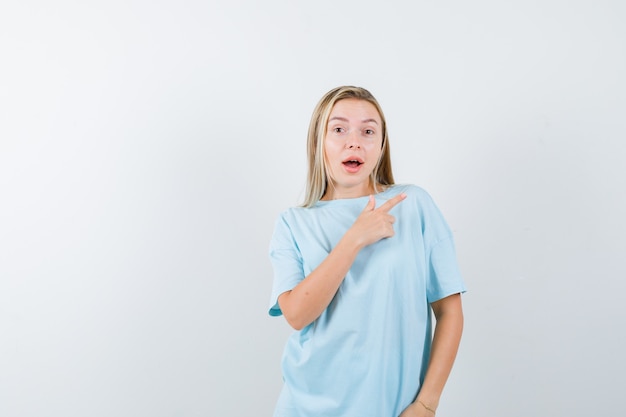 Blondes Mädchen im blauen T-Shirt, das rechts mit Zeigefinger zeigt und überrascht, Vorderansicht schaut.