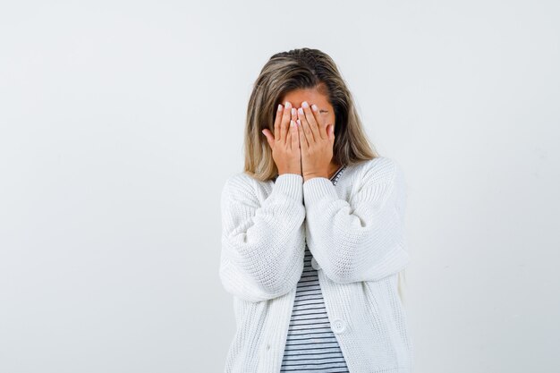 Blondes Mädchen im gestreiften T-Shirt, in der weißen Strickjacke und in der Jeanshose, die Gesicht mit Händen bedeckt und schüchtern schaut, Vorderansicht.