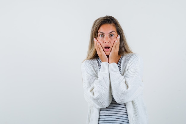 Blondes Mädchen im gestreiften T-Shirt, in der weißen Strickjacke und in der Jeanshose, die Hände auf Wangen halten und überrascht, Vorderansicht schauen.