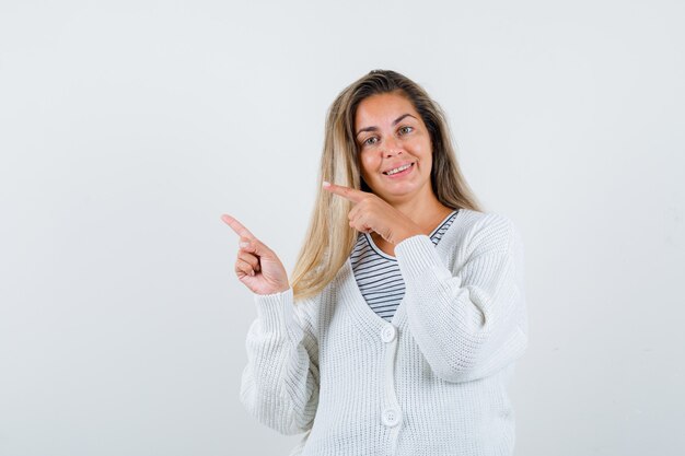 Blondes Mädchen im gestreiften T-Shirt, in der weißen Strickjacke und in der Jeanshose, die mit Zeigefingern nach links zeigen und glücklich schauen, Vorderansicht.