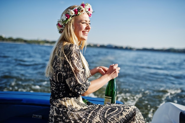 Blondes Mädchen im Kranz sitzt auf Jacht beim Junggesellinnenabschied und öffnet Champagner