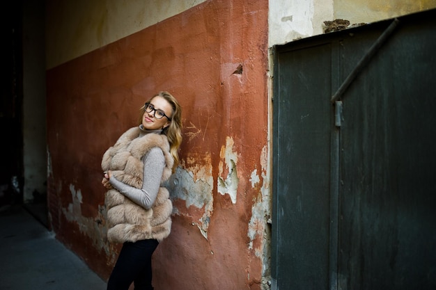 Blondes Mädchen im Pelzmantel und Brille posierte gegen alte orangefarbene Wand