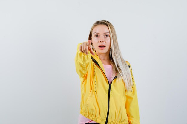 Blondes Mädchen im T-Shirt, Jacke, die auf die Kamera zeigt und erstaunt aussieht