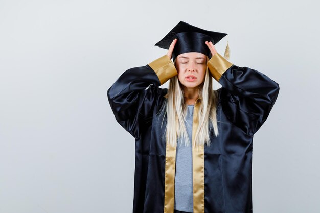 Blondes Mädchen in Abschlusskleid und Mütze, das die Hände auf die Mütze legt, die Augen schließt und ruhig aussieht