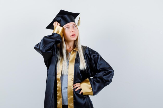 Blondes Mädchen in Abschlusskleid und Mütze, das die Hand an der Taille hält, die Hand auf den Kopf legt, wegschaut und selbstbewusst aussieht